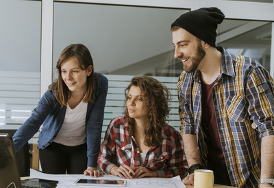 Foto: ein kleines Team arbeitet vor einem Computer an einem Projekt. Auf dem Tisch liegen Papierpläne.